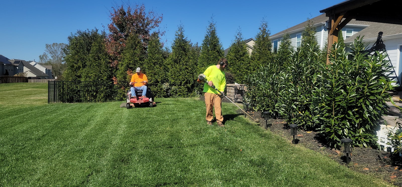 Crew member mid-pass with a walk-behind mower on a residential side yard.